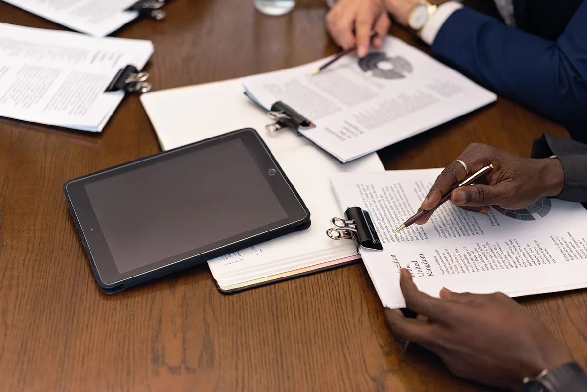 Two people at a table annotating printed documents held in clipboards, a tablet beside them.