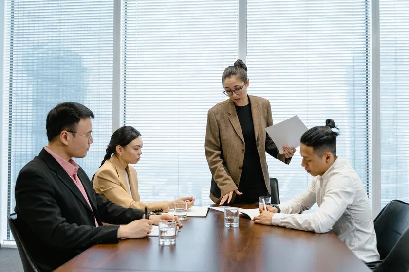 A facilitator standing at a meeting table holding a document while three seated colleagues follow along.